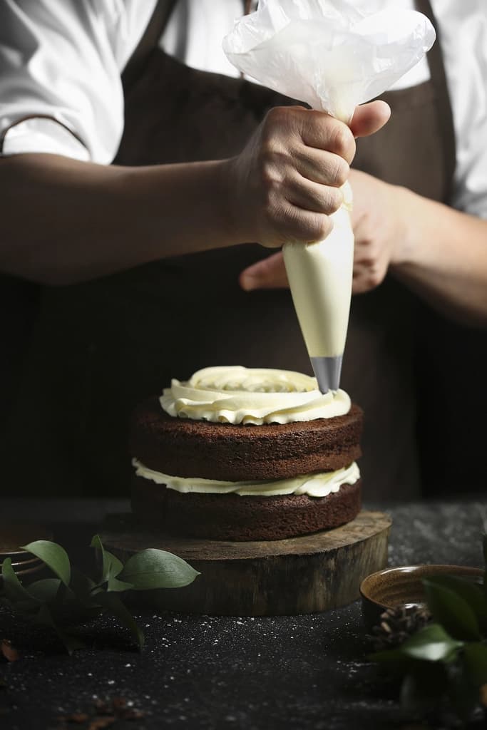 Manos aplicando crema con manga pastelera sobre una tarta de chocolate