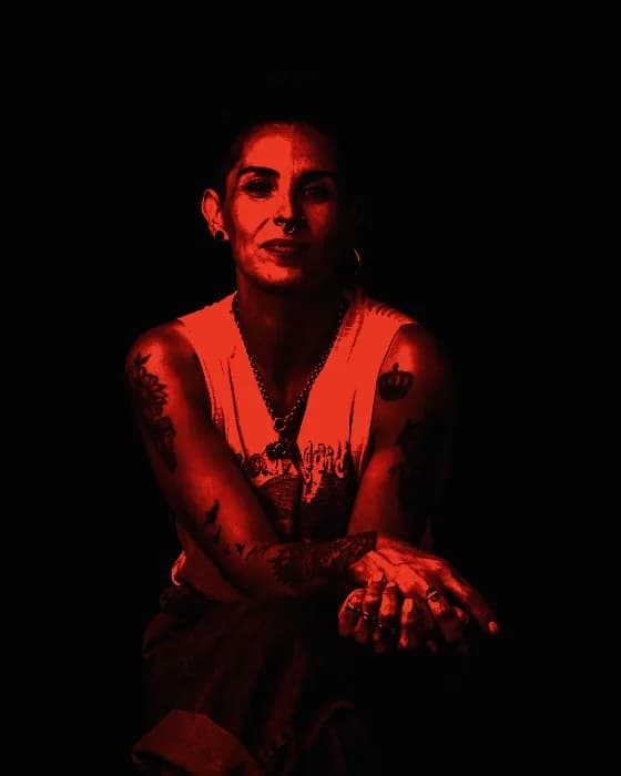 Retrato de Fer Aguirre sentada sobre fondo negro, brazos tatuados y manos cruzadas sobre el regazo
