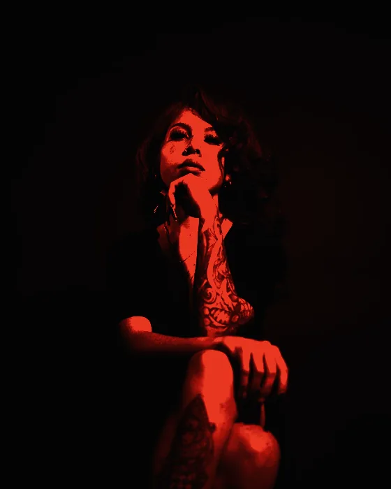 Retrato de Lai Ferrán sentada sobre fondo negro, apoyando la barbilla en la mano, con el brazo tatuado a la vista