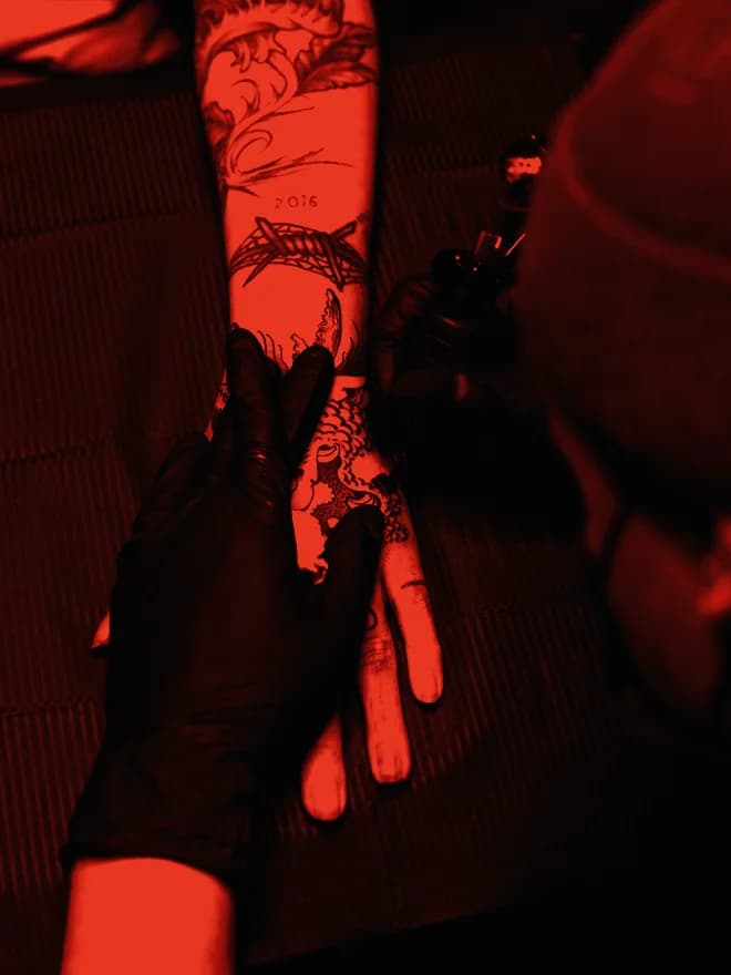Tatuador con gorra inclinado sobre el brazo de un cliente durante la sesión